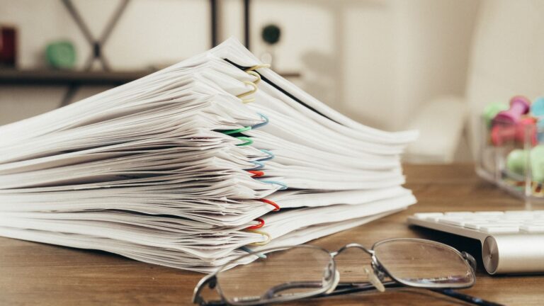 Stack of clipped paper documents and reading glasses on a desk